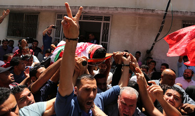 Man makes a V symbol with his right hand while Muhammad Bakr's shrouded body is carried on a stretcher in a crowd of people