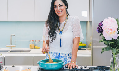 Smiling young woman seen waist-up standing behind kitchen counter while stirring pot on a stovetop