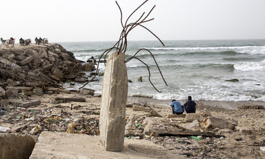 Photo shows the backs of two youths sitting on a beach strewn with litter and rubble