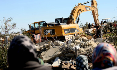 Photo shows the backs of women's heads as they watch a Caterpillar machine being used to raze a home