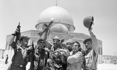 Soldiers seen from waist up raise rifles and helmets above their head while standing in front of Dome of the Rock