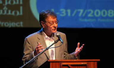 Seen from waist up, Mahmoud Darwish gestures with his hands as he stands behind a podium on a stage