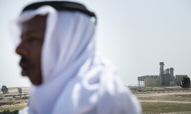 Man wearing head wrap stands in open land in front of power plant