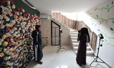 Woman wearing niqab takes a photo of a smiling woman standing in front of a wall decorated with paper flowers