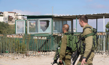 Two soldiers carrying heavy weaponry walk in front of checkpoint with shattered windows