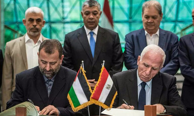 Two men sitting at a desk sign documents as three men standing behind them look on