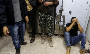 Photo shows mourning man sitting on the ground with his head in his hands as three men, one of them carrying a rifle, standing nearby are shown from waist down