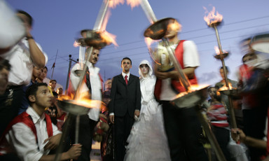 A bride and groom walk through a procession of men in traditional dress carrying burning torches