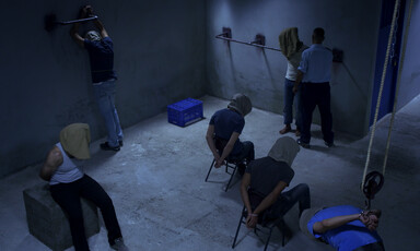 Six men wearing hoods in a dark room are put in various stress positions while a seventh man wearing a guard's uniform stands next to one of them