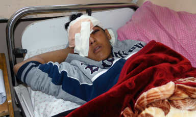 Boy with bandaged head and eye lies on a bed, covered with a blanket