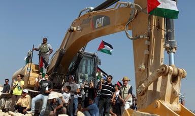 A multigenerational group of people, some carrying Palestine flags, sit around an earth-moving machine with Caterpillar's logo on it and an Israeli soldier standing on top of it