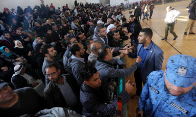 Dozens of people sit in the bleachers of a gymnasium used as a waiting area