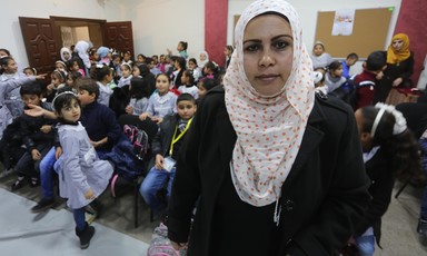 A young woman looking at the camera stands in front of a crowded hall of schoolchildren
