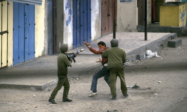 A young Palestinian man gestures towards an Israeli soldier cocking his rifle while being grabbed at by another soldier on the street