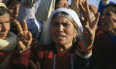 Elderly woman wearing traditional embroidered dress gives victory hand sign among crowd of women