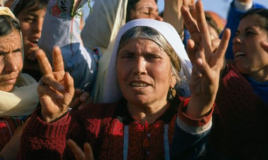 Elderly woman wearing traditional embroidered dress gives victory hand sign among crowd of women