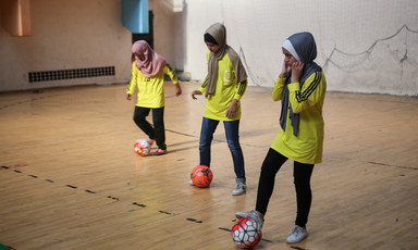 Three girls manipulate footballs with their feet in a gymnasium
