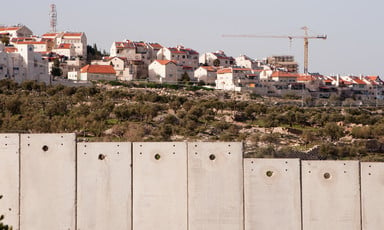Landscape photo shows a construction crane is above a group of Israeli settlement homes with Israel's concrete wall in the foreground
