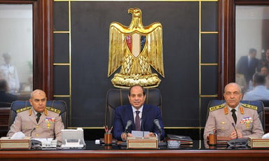 Abdulfattah al-Sisi sits at a desk underneath golden eagle medallion with a military officer on either side of him