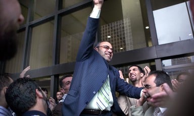 Man in middle of crowd smiles while making victory hand gesture outside court building