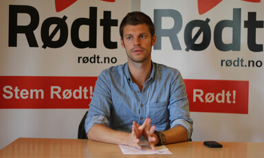 Young man speaks while sitting at desk with a Rødt party banner behind him