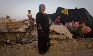 A woman wearing a long black dress stands in foreground as children sit on the rubble of a razed structure in the background