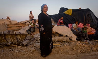 A woman wearing a long black dress stands in foreground as children sit on the rubble of a razed structure in the background