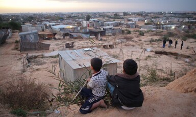 Two boys sit on a sand embankment overlooking a village of corrugated metal shelters