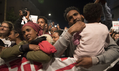 Man holding toddler and woman stand with their arms folded over protest banner