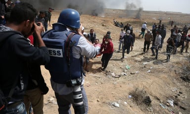 Photographer wearing flak jacket marked as PRESS and wearing a blue helmet is seen from behind while taking photos of demonstrators