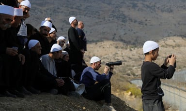 A group of men and boys wearing traditional white caps stand and sit on hillside looking over boundary fence, some holding binoculars