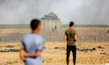 The backs of two standing youth are seen in foreground of photo with Israeli military installation behind barbed wire and fencing in background