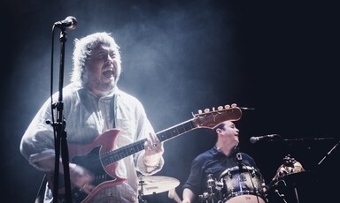 Concert photo of Richard Dawson playing guitar on stage with drummer in background