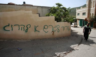 A Palestinian man stands next to a house vandalized with Hebrew-language graffiti. 