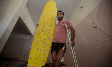 Man wearing t-shirt and shorts holds a surfboard with one arm and supports himself with crutches using his other arm
