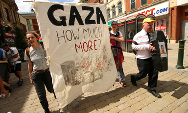 Activists hold large banner reading Gaza How Much More? with image of burning buildings on it