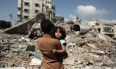 Photo shows back of boy holding small child as the boy stands in front of ruins in urban landscape
