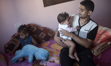 Muhammad al-Najjar smiles and looks at his infant daughter who he is holding while his toddler daughter sits on couch behind him