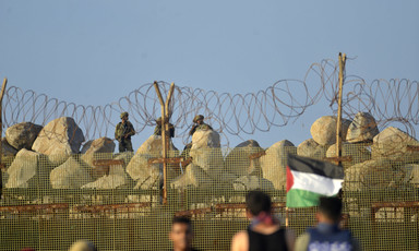 Palestinian protesters stand in front of Israeli soldiers, separated by a barbed wire fence and sandbags