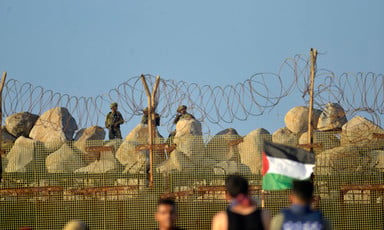 Palestinian protesters stand in front of Israeli soldiers, separated by a barbed wire fence and sandbags