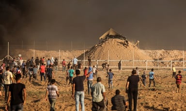 Men and boys stand or run in front of the Gaza boundary fence with an Israeli military installation on a sand hill behind it