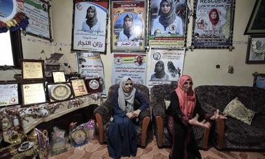 Two women sit on chairs in room with walls covered with posters of Razan al-Najjar in medic's uniform