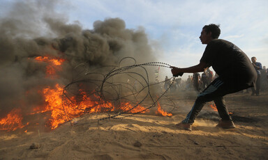 A youth pulls on a barbed wire fence that is partially set on fire as a crowd looks on