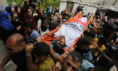 The body of a young man wrapped in a faction flag is carried on a stretcher by a crowd