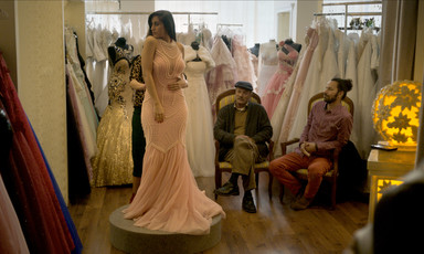 Woman wearing beaded gown stands on pedestal as two men sitting on chairs look on in bridal dress shop