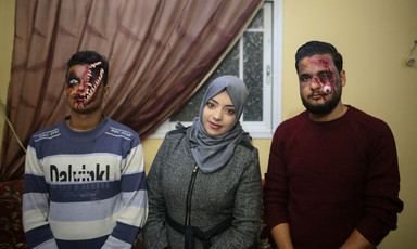 Alaa Abu Mustafa stands between two of her "zombies." One her right, a man has his face apaprently split open, with bones protruding. On her left, a man's eye is made out to be dangling down his cheek. 