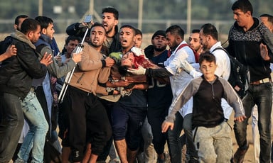 Around a dozen men carry a man who is bleeding from the neck with the Gaza boundary fence in the background