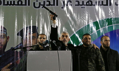 Yahya Sinwar stands behind a podium flanked by two security guards while holding a pistol in the air