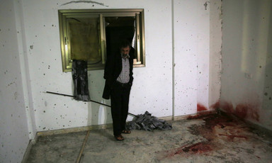 A man stands in front of a window in a room with bullet holes in the walls and a large blood stain in one corner