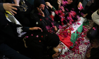 Women lean over and throw flower petals on body of boy shrouded in Palestinian flag